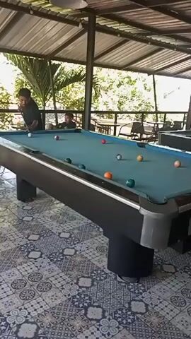 The video shows a group of people playing pool in an outdoor covered area. There are at least two pool tables visible, one in the foreground and another in the background. Several billiard balls are scattered across the table in the foreground. Two individuals are actively engaged in playing, with one person, dressed in a black shirt, positioned at the head of the table, cue stick in hand, seemingly preparing to take a shot. Another person, wearing a green shirt, is standing near the second pool table in the background, holding a cue stick. The environment appears to be a recreational space with a roof overhead, possibly a patio or an open-air establishment. The floor is tiled with a patterned design. Lush green foliage is visible in the background, suggesting a tropical or natural setting. The lighting is natural, indicating daylight.