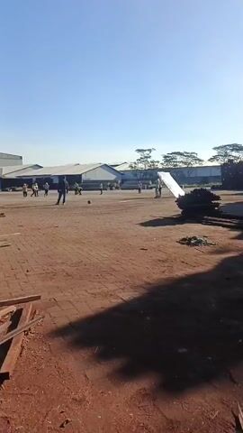 A group of people, some appearing to be men and others women wearing headscarves, are gathered in an open, dusty area. A few individuals are standing and walking around, while many are seated on the ground. In the background, there are several low-lying buildings with long roofs, and a stack of dark, rectangular objects. The ground is a reddish-brown, earthy surface with some scattered debris. Sunlight casts long shadows across the scene, indicating it is likely morning or late afternoon. The sky is clear and blue. One individual, a man in a blue shirt and dark pants, is walking towards the seated group. Another individual, on the left side of the frame, is kicking a ball. A large, reflective surface, possibly a board or a sheet of metal, is leaning against something in the middle ground. The overall impression is of an outdoor gathering or activity taking place in an industrial or undeveloped area.