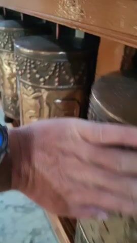 A person's hand is shown in close-up, gently touching and spinning a series of large, ornate, brass prayer wheels. The prayer wheels are cylindrical and decorated with intricate carvings. They are arranged in a row on a wooden shelf. The background is slightly blurred, showing more prayer wheels and hints of the surrounding environment. The video focuses on the texture and movement of the prayer wheels as they are rotated.