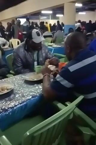 The video shows a crowded dining hall or cafeteria. People are seated at tables covered with plastic tablecloths, eating from plates and cups. Most appear to be adults, and their attire ranges from casual to business-casual. Some are wearing badges or lanyards. The tables are arranged in rows, and the room appears to be well-lit. Individuals are engaged in eating and conversing with each other.