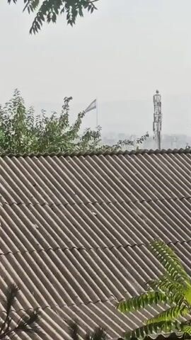 A flag flies on a pole in the distance, possibly the Indian flag. Next to it is a cell tower. In the foreground is a corrugated roof and some trees.