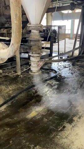 The media file shows an industrial setting, likely a factory or processing plant, with a focus on a piece of machinery. A large, conical hopper, coated in dust and debris, is suspended above a series of rotating augers or feeders. Below this assembly, a white pipe with a flange is positioned, and from it, a spray of white mist or powder is being discharged onto the floor. The floor itself appears to be made of brick or tile, covered in a layer of dark, wet residue and scattered chunks of material. Black hoses snake across the floor, one of which appears to be supplying the mist. In the background, more industrial equipment, pipes, and structural elements are visible, suggesting a large, possibly disused or operational facility. The overall impression is one of a messy, working environment, with evidence of material being processed or cleaned. There is no indication of the time of day, weather, or specific location beyond the general industrial context. The limited visibility of the machinery and the focus on the discharge suggest a process-related activity, perhaps related to material transfer, dust suppression, or cleaning. There is no visible text in the media file.