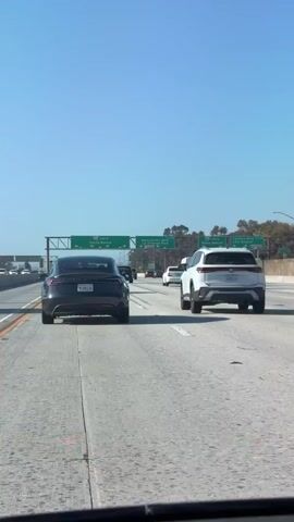 The video shows a first-person view from inside a car driving on a highway. The car's dashboard displays a speed of 66 mph, with the speed limit set at 55 mph. Multiple cars are visible on the highway, including a white SUV, a black sedan, and other vehicles in the distance. Overhead green highway signs indicate upcoming exits for Santa Monica Blvd and Manchester Blvd. The sky is clear and blue. The overall environment appears to be a sunny day on a busy highway.