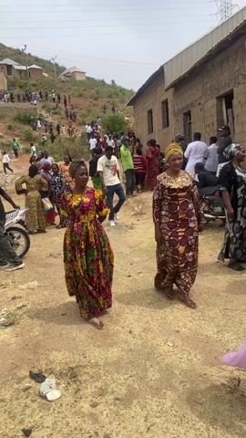 The video captures a large gathering of people in an outdoor, semi-urban or rural setting. The ground is mostly dry, light-colored dirt, with some patchy vegetation. In the foreground, several individuals are walking along a dirt path. Two women are prominent; one on the left wears a vibrant, multicolored floral dress, while the other on the right is dressed in a dark brown and gold patterned gown. Many other women in the crowd are also seen in similar patterned dresses, often in bright colors. Men are dressed in casual wear, including t-shirts and trousers. One man, walking behind the foreground figures, wears a white t-shirt with the word "STRAIGHT" visible on the front.

In the mid-ground and background, the landscape rises steeply into a hill, which is also populated with many people. Scattered across the scene are simple, unpainted buildings, some appearing to be partially constructed with exposed concrete blocks. A motorcycle with the brand name "JINCHENG" visible on its side is parked near the left edge of the frame. The overall impression is one of a bustling community event or gathering, with people mingling, walking, and standing in groups. The weather appears to be clear and sunny. The ambient sound includes indistinct human voices and chatter.