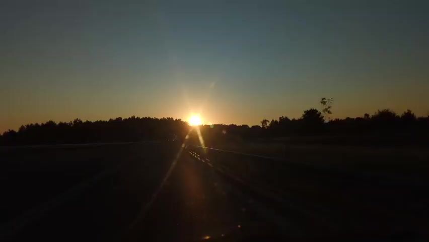 The video shows a point of view from inside a moving vehicle on a road during sunset. The sun is low on the horizon, casting a warm glow and creating lens flares that are visible on the windshield. The road has white double lines, and there is a forest or treeline visible on both sides of the road. A truck can be seen in the distance on the left side of the road. The sky is a gradient of orange, yellow, and blue, indicating the time of day. The overall impression is that of a journey at dusk.