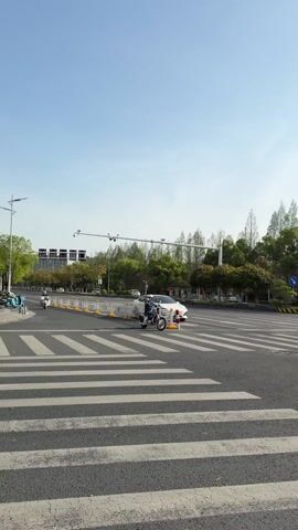 The video shows a crosswalk in a city or suburban area. In the first few seconds, a person on a scooter crosses the crosswalk from left to right. A white car is stopped nearby. After the scooter passes, the camera pans to the right, revealing a green truck parked on the side of the road. There are trees and buildings in the background. The sky is blue with the sun shining.