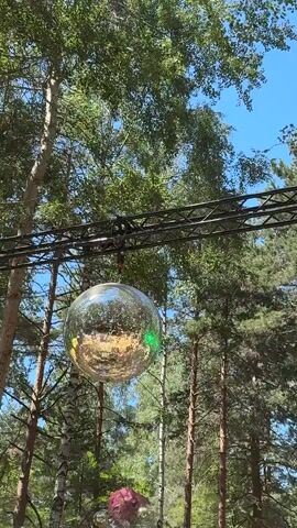 The video begins with a close-up of a disco ball hanging from a metal truss structure in a wooded area. The disco ball is reflecting light and surrounded by greenery and blue sky. The camera then pans to the right, revealing more trees and a glimpse of a keyboard covered with a purple cloth, placed on a stage-like structure. Smoke is present around the keyboard. The scene shifts to a DJ booth with a DJ at the turntables. Bubbles are floating in the air around the DJ booth and in front of a stage structure. The DJ is wearing a patterned shirt and appears to be operating the equipment. The environment is outdoors, surrounded by trees, suggesting an event or festival setting.