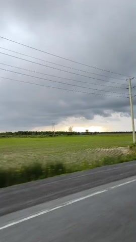 This media file captures a dynamic landscape view from a moving vehicle, likely a car, traveling along a paved road. The road is in the foreground, with a white line marking its edge. Beyond the road, a vast, vibrant green field stretches towards a distant tree line. Power lines crisscross the sky, supported by utility poles visible in the frame.

The sky is dramatic, filled with heavy, gray clouds that suggest an impending storm or recent rainfall. A sliver of sunlight breaks through the clouds on the horizon, casting a soft glow over the distant landscape and hinting at either sunrise or sunset. The weather appears to be overcast and potentially stormy, contributing to a sense of atmospheric intensity.

No people are visible in the media file. The primary subjects are the expansive agricultural field, the infrastructure of power lines and poles, and the ever-changing sky. The setting is rural, characterized by open fields and the absence of urban development. The motion blur on the field indicates the speed at which the recording is taking place. The overall mood evoked is one of natural beauty combined with a sense of raw, untamed weather. The location is specified as Champlain, Canada. No text is visible in the media file.