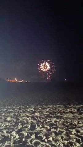 The video shows a nighttime scene on a sandy beach. In the distance, fireworks are being set off. The fireworks are a mix of colors, primarily white and pink, and explode in a circular pattern. The beach in the foreground is dark, with the texture of sand visible. A faint line of lights from a distant town or boardwalk can be seen along the horizon. The sky is dark and clear, with no visible moon.