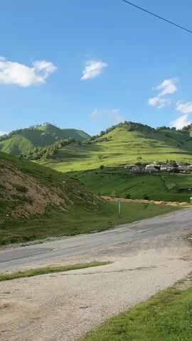The video shows a rural landscape with a paved road winding through rolling green hills under a blue sky with scattered white clouds. In the distance, a small village is nestled among the hills, with houses and a church visible. The foreground is dominated by the road and the grassy slopes on either side. A utility pole with wires stretches across the frame. The camera pans slowly from left to right, revealing more of the village and the surrounding terrain. The sun is shining, casting shadows across the landscape, suggesting it is either morning or late afternoon. The overall impression is one of tranquility and natural beauty.