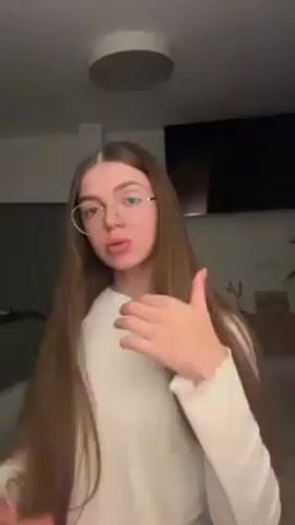 A woman with long, straight, light brown hair and glasses is standing in a room. She's wearing a white, long-sleeved shirt. She moves her hair with her hands, then touches her shirt collar. She then poses slightly, tilting her head and pursing her lips. The background includes a ceiling light, a TV, and furniture.
