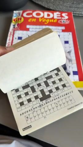 The video shows a person flipping through the pages of a puzzle book, revealing completed codeword puzzles. The book has a black and white grid layout with filled-in letters in black and blue ink, and purple ink on a later page. A handwritten key associating numbers with letters is visible below each puzzle grid. A "CODES en Vogue" magazine is in the background, featuring a crossword puzzle and an image of a white horse on its cover. The surface the puzzle book is lying on is a light gray color, possibly a table.