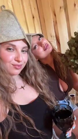 Two young women are in a sauna, as indicated by the wooden walls and their sauna hats. The woman on the left is wearing a black tank top and a necklace with a cross pendant. She's holding a dark-colored drink in a goblet and smiling at the camera. The woman on the right is wearing a brown top and is holding a bundle of green leaves. They appear to be enjoying themselves in the sauna.