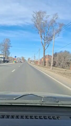 The video shows a vehicle driving down a straight, two-lane road on a sunny day. The sky is blue with scattered clouds. Houses are visible on the right side of the road, and some trees line the roadside. Streetlights and power lines run along the road. Other vehicles, including trucks, are visible in the distance. There is a road sign on the left side of the road. The interior of the car, specifically the dashboard, is visible at the bottom of the frame.