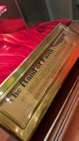 The video shows a display case featuring "The Hand of Faith" gold nugget. The camera pans up from a gold-framed information plaque to reveal the nugget itself, displayed within a clear, hexagonal case atop a red velvet cushion. Two similar cases containing other gold nuggets are also visible behind the main nugget, also resting on red velvet cushions. The background appears to be a well-lit exhibit area.