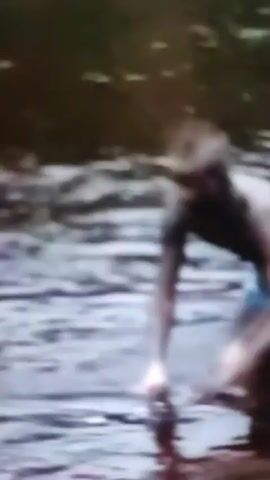 A person is in the water, partially submerged. The person appears to be struggling or swimming. The water is choppy and dark. The surrounding environment is out of focus but appears to be natural.