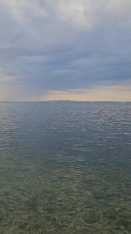 The camera shows a view of a body of water. In the distance, many small white objects, potentially sailboats, are visible against a slightly lighter horizon. The water near the camera is clear enough to see the pebbles beneath the surface. The sky is overcast with grey clouds. On the left, there is a small land formation with sparse tree cover.