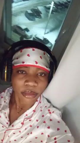 A person wearing a white pajama top with red hearts, a matching head scarf, and headphones looks directly into the camera. The background shows a window with a view of a parking lot with cars covered in snow. The person appears to be indoors, possibly leaning against a wall near the window. The lighting suggests it is nighttime or a dimly lit indoor space.