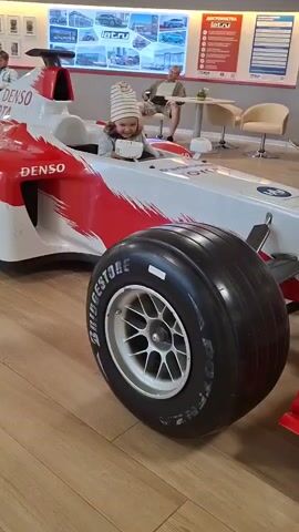 A young child, wearing a striped beanie and a grey long-sleeved shirt with a printed design, sits in the driver's seat of a white and red Formula 1 racing car toy. The child is holding onto the steering wheel and looking forward. The car has the "DENSO" and "TOYOTA" logos visible on its side. A large tire with "BRIDGESTONE" written on it is in the foreground. In the background, a man is sitting at a table, and a wall display with images and text is visible. The flooring appears to be wood laminate. The lighting is bright and indoor.