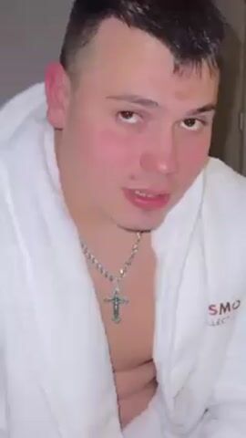 A fair-skinned young man with short, dark hair, red cheeks, and a silver cross necklace is seen wearing a white terrycloth bathrobe. He appears to make a puffing-out sound and then smirks slightly. The robe obscures the background which appears to be a doorway. The letters "SMO" are visible on the robe.