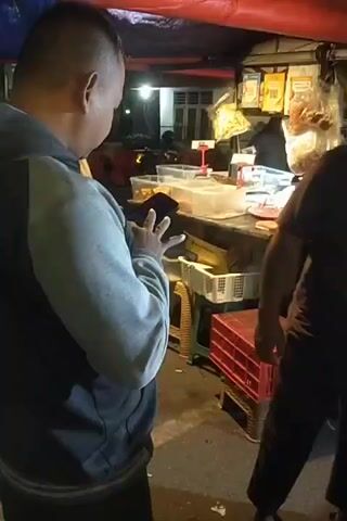 The video shows an outdoor scene at night, likely a street market or food stall area. In the foreground, two men are interacting. One man, facing away from the camera, is wearing a grey hooded jacket and appears to be looking at a mobile phone. The other man, facing towards the camera, is wearing a dark t-shirt with a graphic design and is smoking a cigarette. He is positioned to the right of the first man. In the background, there are more people and what appear to be food stalls or vendor tents, illuminated by artificial lights. A woman in a hijab and long skirt is visible standing near a stall. Other figures and structures suggest a bustling outdoor environment. The lighting is dim, typical of nighttime street scenes.
