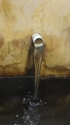 A close-up shot shows water pouring from a white pipe. The pipe is affixed to a tan, stained wall. The water is flowing into a dark, murky basin, creating ripples and splashes on the surface.