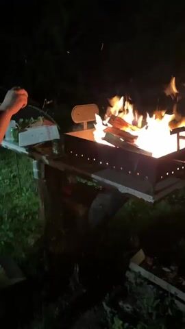 The video opens with a close-up of a metal brazier containing a large, active fire with bright orange flames and upward-flying sparks, positioned outdoors at night. The brazier sits on a wooden stand. A young, light-skinned male with short brown hair, wearing a light-colored, short-sleeved collared shirt, is then shown holding a black hose-like object with both hands, swinging it back and forth with effort and a smile. He is positioned to the left of the brazier, which is visible with the fire still burning. A brief glimpse of another person's arm is seen behind him. The video concludes with another shot of the brazier and the burning fire, with the surrounding area illuminated by the flames and the night environment in the background.