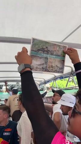 A group of tourists on a boat are looking at a laminated picture of a cave labeled "Viking Cave". The picture shows four different images of the cave's entrance and interior. The tourists are on a boat with a white canopy, and the water is visible around them. Some people are wearing hats and sunglasses. One person in the foreground is wearing a black shirt and arm sleeve, holding the picture up for others to see. Another person is wearing a white bucket hat and an orange shirt. The video appears to be taken during the daytime.