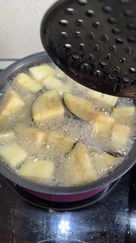 A person is cooking cut-up eggplants in a pot with boiling water. They are using a black slotted spoon to stir the eggplant pieces in the pot. The pot is on a black electric stovetop. The eggplants are mostly submerged in the water.