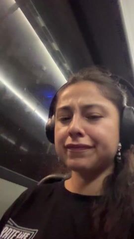 A woman is wearing headphones and sitting in a moving vehicle, likely a bus or train. She is wearing a black long-sleeved shirt. It is raining outside, and the rain can be seen on the window. The woman clenches her fist and moves it up and down, and then smiles. The lighting in the vehicle appears to be fluorescent.