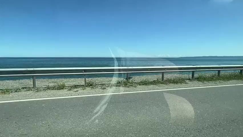 A car travels along a road next to the ocean. The sky is blue and clear. The ocean is calm and blue. There is a guardrail between the road and the ocean. Grass and some rocks are visible on the side of the road. The view is from inside the car, looking out the window. The side mirror of the car is visible on the right side of the frame.