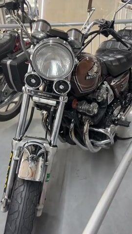 A 20-second video shows a close-up view of a vintage Honda motorcycle parked indoors. The motorcycle is dark brown with chrome accents. The video pans from the front of the motorcycle, showing the headlight and front forks, to the seat and the side of the engine. The Honda logo is visible on the fuel tank. The background appears to be a workshop or showroom, with metal barriers and other objects blurred in the distance. There are no people visible in the video.