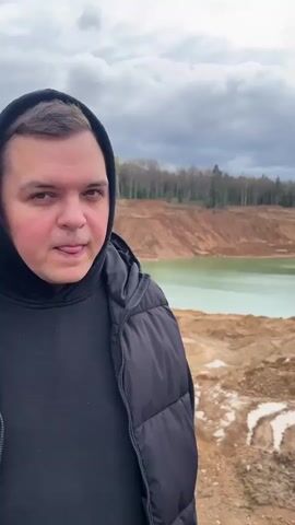 A man in a black hooded jacket stands in front of a clay pit with a pool of turquoise water in the background. He makes a "V" sign with his fingers and smiles at the camera. The sky is cloudy and overcast. There are trees in the far background.