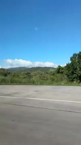The video shows a view from a moving vehicle driving down a highway. The sky is bright blue with a few scattered clouds. On the right side of the road, there's a grassy bank covered with tall green plants and some trees. In the background, there are green hills or mountains. A white line marks the edge of the road. A highway sign with the number "991" is visible. The video appears to be taken on a sunny day.