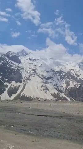 The video shows a snow-covered mountain range under a blue sky with scattered clouds. The foreground consists of a dirt or gravel ground. There are no visible people or animals. The camera appears to be static.  Background music is audible.