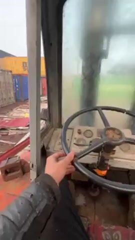 A person sits inside the cabin of a large machine, possibly a crane or forklift, with a steering wheel in their hand. The view from the machine's window shows an outdoor area with shipping containers of various colors scattered around. There is debris on the ground, including metal sheets and other discarded materials. The environment appears to be a storage or salvage yard. It is a hazy day, with a muted overcast sky. The person seems to be either starting or maneuvering the machine, judging by the sounds and slight movements.