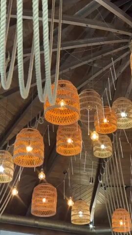 The camera pans from the ceiling of a circular wooden room with numerous woven lantern-style light fixtures hanging from it, to a long wooden table surrounded by people. The ceiling is adorned with a pattern of ropes. Three women and a man sit around the table, which is set with water bottles, glasses, and floral arrangements. The woman in the foreground on the left looks directly at the camera. The man next to her wears sunglasses and leans back in his chair.