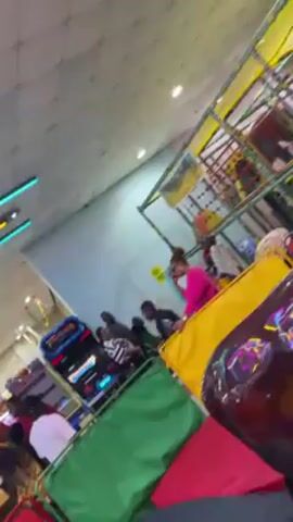 The video pans across a colorful arcade, highlighting the neon lighting and various game machines. There are several people of different ages present, some playing games and others walking around. The camera focuses briefly on a young girl smiling at the camera. The overall atmosphere appears lively and playful.
