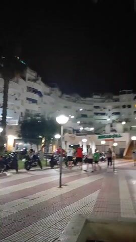 A night scene shows a curved white building with multiple balconies and windows, some illuminated. Palm trees are visible in front of the building. A sidewalk with a red and white striped pattern runs in the foreground, with streetlights along it. Several motorcycles are parked along the sidewalk. A group of people are standing and walking on the sidewalk, some appearing to be interacting. One person is walking towards the camera. The sky is dark, indicating nighttime. A sign for "Mercadona" is visible on the building.