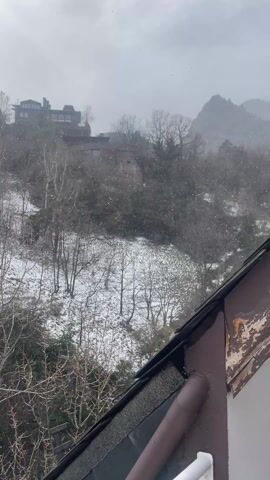 The video shows a view from what appears to be a roof or elevated balcony, looking out over a wintry landscape. Snow is falling lightly, and there is a dusting of snow on the ground, trees, and rooftops. The foreground includes a section of the roof and a gutter. The middle ground features a wooded area with bare trees and some evergreen trees. In the background, there are buildings situated on a hill, partially obscured by the falling snow and distant landscape. The overall impression is a quiet, snowy winter scene.