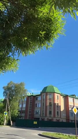 The video begins with a close-up of lush green tree branches against a bright blue sky. The camera pans down to reveal a multi-story building with a distinctive green, octagonal roof and red brick accents. A street with parked cars is visible in the foreground, and a traffic sign for pedestrian crossing is present. The scene transitions to show more apartment buildings and trees, with parked cars lining the street. The sky remains clear and blue with some fluffy white clouds. The sun is visible, casting a bright light on the scene. The overall impression is an urban residential area on a sunny day.