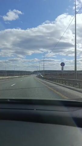 The video shows a view from inside a vehicle traveling on a highway. The road is asphalt, marked with a yellow line, and lined with streetlights and metal guardrails. Beyond the guardrails, there is a field and some trees. The sky is blue with scattered white clouds. The dashboard of the vehicle is visible at the bottom of the frame.