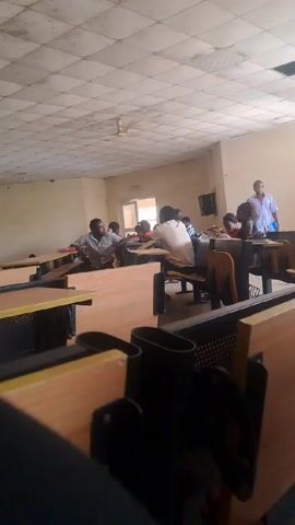 The scene appears to be inside a classroom with a tiled ceiling and fluorescent lighting.  There are rows of wooden desks with black metal frames and attached seats. Several individuals are sitting at the desks, some interacting with each other.  One man wearing a light-colored shirt is standing and appears to be talking or addressing the others.  A window is visible on the far wall and there are a few other details like a small television screen also on the wall. The perspective seems to be from the back of the classroom, between the rows of desks.