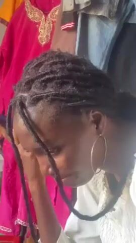 A young woman with dark skin and dreadlocks is looking down and holding a purple object near her face. She is wearing large gold hoop earrings and a white shirt with a floral pattern. Behind her is a pink garment with gold embroidery. Other clothing items are also visible in the background.