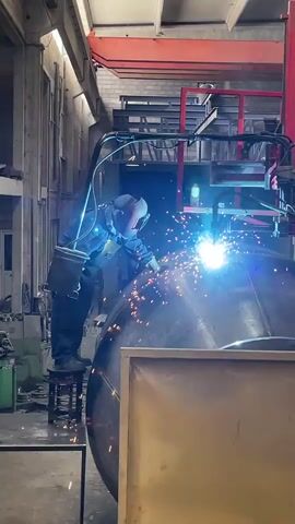 A welder in full protective gear, including a helmet and dark blue overalls, stands on a stool next to a large, cylindrical metal tank. The welder is actively welding the tank, indicated by the bright arc of light and a shower of sparks emanating from the point of contact. The welding equipment, including a hose and a power source, is visible. The environment appears to be a workshop or factory, with industrial machinery and structures in the background, including a red overhead crane. The lighting is dim, with the bright welding arc being the primary light source in the immediate vicinity.