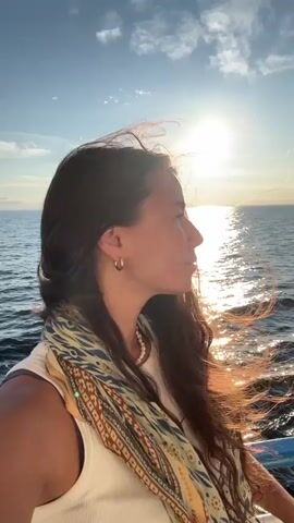 A medium shot video shows a young woman from the chest up, looking out at the sea during sunset. Her long, dark hair is blowing in the wind, partially obscuring her face. She is wearing a white tank top and a patterned scarf draped around her neck. The sun is setting behind her, casting a bright glow and reflecting on the water. The sky is blue with some scattered clouds. The video appears to be shot on a boat, with the railing of the boat visible at the bottom of the frame.
