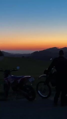 The video opens with a shot of two dirt bikes parked side-by-side on a paved surface. A person, silhouetted against a vibrant sunset sky, stands between the bikes, looking out over a hilly landscape. The sky transitions from a deep blue at the top to orange and pink hues near the horizon. The landscape features rolling hills and fields, with trees dotting the scene. As the video progresses, the image briefly blurs, showing a dark expanse, likely trees or a forest. The final shot shows a full moon hanging in a clear blue night sky above a small village nestled in the hills. Tall, dry grass fills the foreground, and the lights of the village are visible in the distance. The overall tone is peaceful and scenic, capturing the transition from sunset to night in a rural setting.