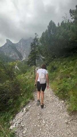 A woman with a white backpack and white t-shirt walks up a rocky path in a forest with mountains in the background. The path is lined with green bushes and trees on both sides. The sky is overcast and cloudy.