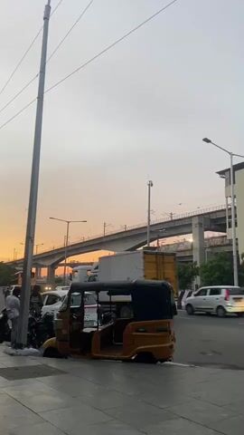 The video captures a street scene during what appears to be either dawn or dusk, indicated by the warm, orange-hued sky. In the foreground, on the left side of the frame, there is a gray sidewalk with a tall utility pole. Adjacent to the sidewalk, a yellow auto-rickshaw is parked, its driver's side door open and its body resting low on what seems to be a flat tire or a significant suspension issue. Several motorcycles are parked behind the auto-rickshaw, and a few individuals are standing near them, seemingly engaged in conversation or waiting.

The street itself is paved and shows signs of traffic. Several vehicles are visible: a white car, a truck with a yellow container, and another white SUV further down the road. A red car passes by in the middle distance.

In the background, an elevated structure, likely a highway or a metro line, spans across the frame, supported by concrete pillars. The sky above is mostly clear with a gradient of orange and pale yellow. To the right of the elevated structure, a modern building with several windows is visible. A street lamp is present on the right side of the road. The overall impression is a busy urban environment at a transitional time of day, with a focus on the parked auto-rickshaw and the surrounding activity.