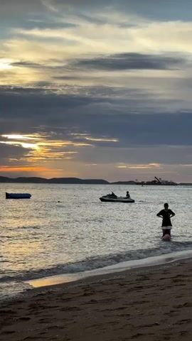 The video shows a beach scene at sunset. The sky is filled with dramatic clouds, with the sun setting behind a distant landmass on the left. The water is calm, with gentle waves lapping at the shore. In the foreground, a sandy beach is visible. In the water, a person is standing near the shore, appearing to be interacting with another person or an object in the water. Further out in the water, a jet ski is visible with at least one person on it. To the left, a blue inflatable raft is floating on the water. In the distance, along the horizon, there are faint structures that suggest a developed area. The overall atmosphere is peaceful and serene, capturing the beauty of a coastal sunset.